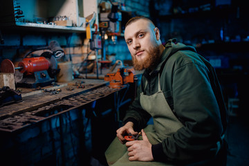 Man with beard in overalls makes details of iron and wood in garage workshop, handmade production....