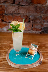 Refreshing cocktail with alcohol, mint, orange and ice in tall glass. Peach is lying on chaise longue on beach.