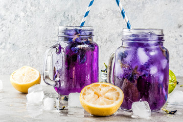 Healthy summer cold beverage, iced organic blue and violet butterfly pea flower tea with limes and lemons, grey concrete background copy space top view
