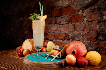 Refreshing cocktail with alcohol, mint, orange and ice in tall glass. Background, mix of fruits: pineapple, kiwi, lemon, pomegranate.