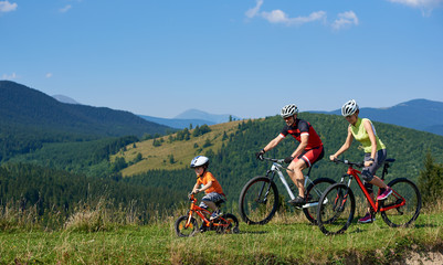 Fototapeta premium Young modern family tourists bikers, mom, dad and child riding on bicycles on grassy hill. Carpathian mountains, blue summer sky on background. Active lifestyle, traveling and happy relations concept.