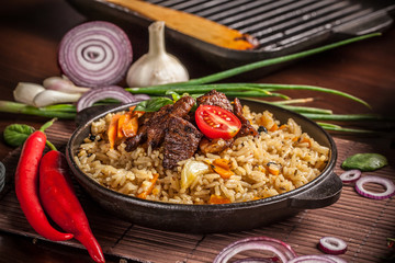 The concept of eastern, Uzbek cuisine. Pilaf, plov in a black cast-iron frying pan on a wooden table, next to lie ingredients, onions, tomatoes, parsley, pepper. Central Asian cuisine.