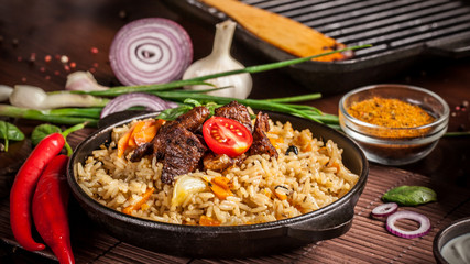 The concept of eastern, Uzbek cuisine. Pilaf, plov in a black cast-iron frying pan on a wooden table, next to lie ingredients, onions, tomatoes, parsley, pepper. Central Asian cuisine.