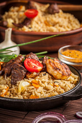 The concept of eastern, Uzbek cuisine. Pilaf, plov in a black cast-iron frying pan on a wooden table, next to lie ingredients, onions, tomatoes, parsley, pepper. Central Asian cuisine.