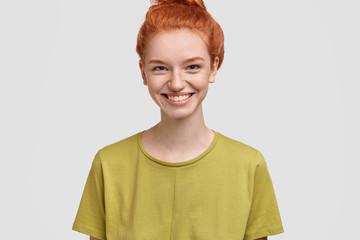 Cropped shot of pleased smiling sensual red haired female has broad smile, white even teeth, dressed in green t shirt, poses against white concrete wall. Attractive woman student recieves good mark