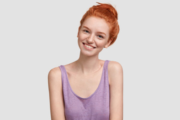 Joyful ginger female with freckled healthy skin, being happy to spend vacation in summer camp, enjoys free time with friends, has red hair, dressed in casual oversized t shirt, isolated on white wall
