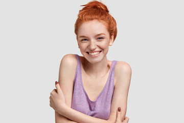Charming lovely ginger female youngster takes part in interesting conversation, has bright smile on face, keeps hands crossed, looks with curiosity at camera, isolated on white studio background