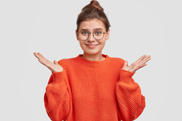 Indifferent pretty young woman shrugs shoulders, wears red sweater, keeps palms raised, poses...
