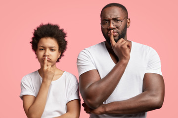 Horizontal shot of handsome dark skinned plump male stands next to his little son, have displeased expressions, discontent with result of football match, keep fore fingers on lips, frown faces