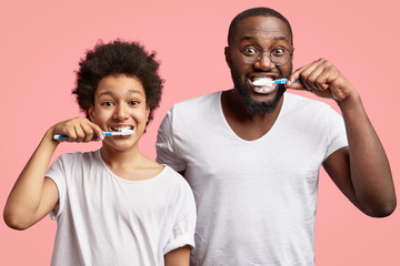 Positve dark skinned son and father brush teeth, keep mouthes wide opened, have satisfied...