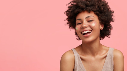 Cute joyful female teenager has broad smile, sparkles on face, prepares to go on disco with friends, being in high spirit, has bare shoulders, stands against pink background, blank space for your text