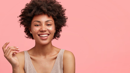 Adorable dark skinned female with glitter on face, smiles broadly at camera, expresses positive emotions, being satisfied with professional make up in beauty salon, isolated on pink background