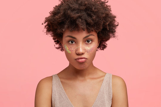 Lovely young female looks seriously at camera, has full lips, dark pure skin and Afro hairstyle, poses against pink background, demonstrates glitter on cheeks, makes photo for fashion magazine