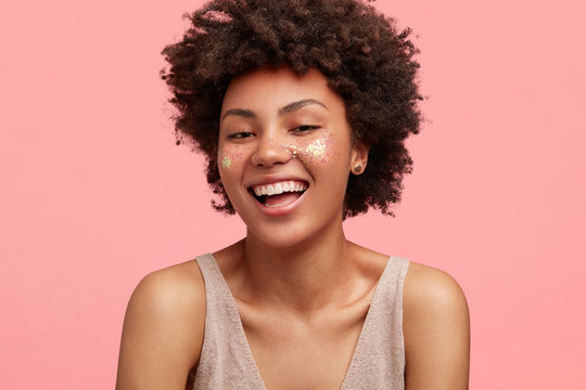 Close up portrait of happy African American female has glitter, broad joyful smile, dressed in casual clothing, rejoices spare time in friendly company, poses against pink background. Beauty concept