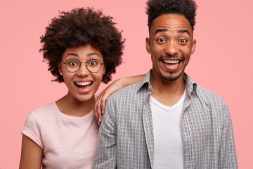 Shot of pleased dark skinned students rejoice successfully passed exam, look positively at camera. Happy African American female feels support from boyfriend, stands closely, isolated on pink wall