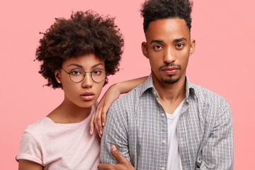 Lovely African American couple stand close to each other, look seriously at camera, have confident expressions, dressed in casual outfit, pose together against pink background. Companionship concept