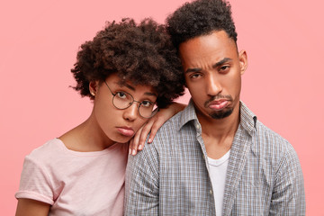 Unhappy African American couple have depressed and sad expressions, have financial problem, feel desperate, purse lips, need support and help, isolated over pink background. Negative emotions