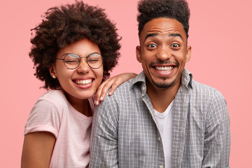 Positive dark skinned female and male being in love, watch comedy, smile happily at camera, laugh at funny scene, enjoy recreation time, isolated over pink background. People, togetherness concept