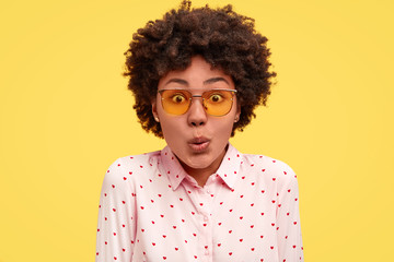 Close up portrait of puzzled young African American female looks in bewilderment, expresses great suprisement, being impressed by something unbelievable, wears blouse, stands against yellow background