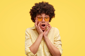 Pretty young African American woman gasps in amazement, keeps mouth wide opened, makes shocking revelation, wears trendy sunglasses, stares at camera, isolated over yellow studio background.