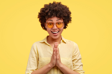 Indoor shot of cheerful pretty African American female keeps palms together, looks friendly and happy, greets someone, shows begging gesture, dressed in blouse, isolated on yellow wall. Monochrome