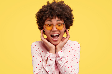 Excited amazed beautiful African American female looks with joyful expression, keeps hands under...