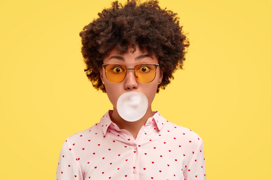 African American female blows bubble from chewing gum, has amazed expression, dressed in fashionable clothes and shades, isolated over yellow background. Lovely dark skinned woman poses indoor