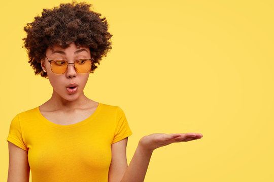 Horizontal shot of amazed fashionable female youngster with dark skin and Afro hairstyle, keeps palm raised as if holds something, advertises blank space wears casual yellow t shirt, bright background