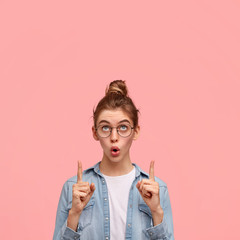 Obraz premium Amazed European female looks upwards with wonderment, indicates with both fingers up, has surprised expression, dressed in fashionable clothing, isolated over pink background with copy space