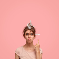 Vertical shot of unhappy beautiful young female with displeased expression, has feather on head,...