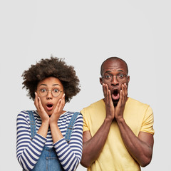 Vertical shot of stupefied African American male and female stare at camera with great surprisement, keep hands on cheeks, realize shocking news, pose against white background. Shock concept