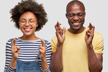 Photo of cheerful African American couple have joyful expressions, cross fingers and smile gladfully, wear casual clothes, isolated over white wall. Happy dark skinned female and male gesture indoor