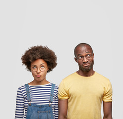 Displeased African American female and male companions curve lips and look with puzzled expression, notice something strange upwards on ceiling, spend free time together, isolated over white wall
