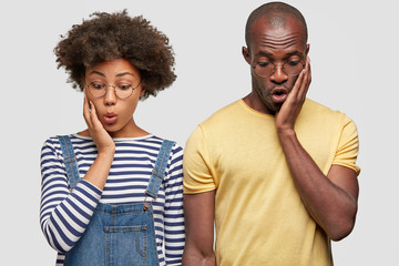 Wow, what is on floor! Surprised young dark skinned male and female couple keep hands on cheeks, look with amazed face expressions down, pose against white background, notice something strange
