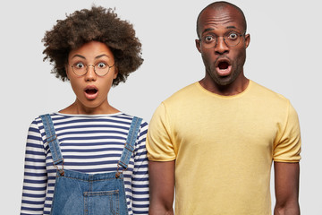 People, emotions and facial expressions concept. Terrified African American female and male see something unbelievable, keep jaws dropped, stand close to each other, isolated over white background