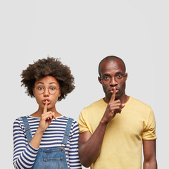 Serious dark skinned female and male keep fingers on mouthes, look secretly at camera, demonstrate silence sign, dressed in casual clothing, isolated over white background. Ethnicity and conspiracy - obrazy, fototapety, plakaty