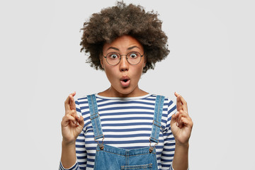 Horizontal shot of amazed beautiful dark skinned female has Afro haircut, crosses fingers, dressed in striped sweater and denim overalls, poses against white background. People and wish concept