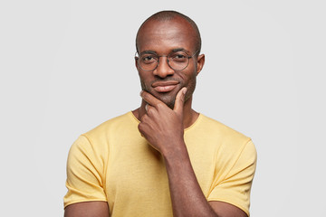 Studio shot of confident pleased African American male boss thinks about new investment plan, holds chin and looks directly at camera, stands against white background. Clever dark skinned guy