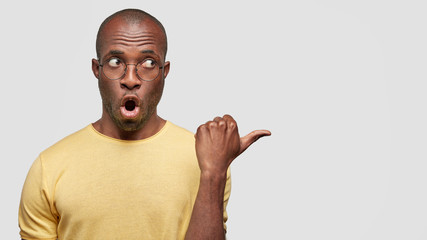Surprised African American male with astonished expression, indicates aside, demonstrates blank space for your advertising content, wears round spectacles, has dark pure healthy skin isolated on white