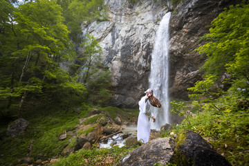 European man with beard is doing waterfall-meditation while standing under big waterfall in...