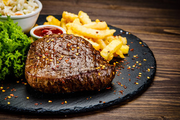 Grilled steak with french fries and vegetables served on black stone on wooden table