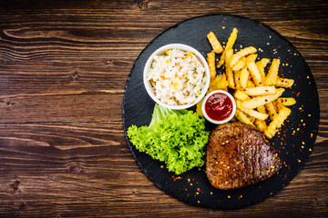 Grilled steak with french fries and vegetables served on black stone on wooden table