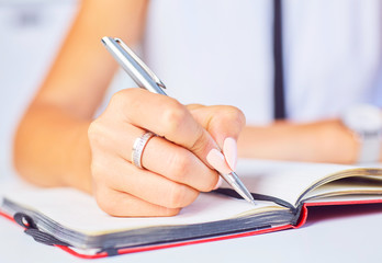 Young woman working at her desk taking notes closeup. Focus on hand writing on a notepad.