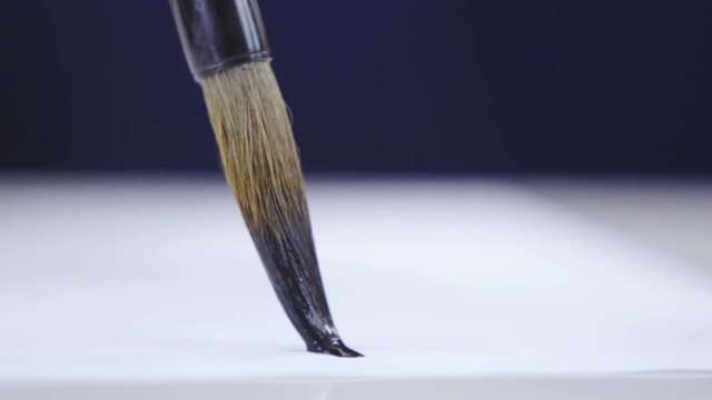 Dipping brush in to black ink and drawing undefined characters 4K. Long shot close up of brush in focus, tracking from ink stone to white paper where brush leaves black mark. 