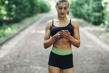 Woman Runner in the Summer Morning Park Listening to Music on Smartphone Using Bluetooth Earphones. Female Fitness Girl Jogging on Path Outside.