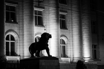 Silhouette of a lion statue against the background of columns of a courthouse. A black and white photo is taken in the evening. Background lighting emphasizes the texture of the columns and brick wall