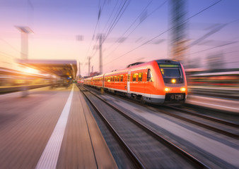 High speed red train with motion blur effect on the railway station at sunset. Landscape. Modern intercity passenger train in motion on the railway platform at dusk. Commuter vehicle on railroad