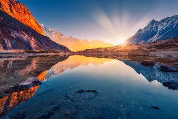 Beautiful landscape with high mountains with illuminated peaks, stones in mountain lake, reflection, blue sky and yellow sunlight in sunrise. Nepal. Amazing scene with Himalayan mountains. Himalayas - obrazy, fototapety, plakaty