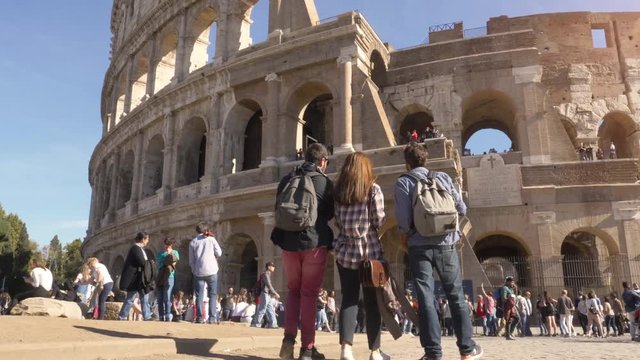 Three young friends tourists standing in front of colosseum in rome with backpacks sunglasses happy beautiful girl long hair