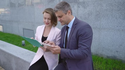 Business man and woman businesspeople sitting on the railing near the office building and discussing the project. A man and a middle-aged woman. people are holding documents and a tablet 4k - Powered by Adobe